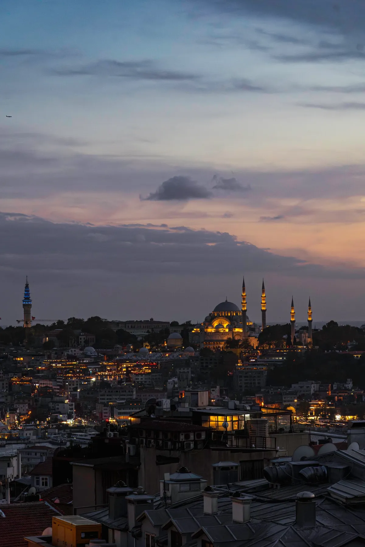 Istanbul rooftops at dusk with a lit mosque and four minarets and a blue-lit tower under a pink sky.