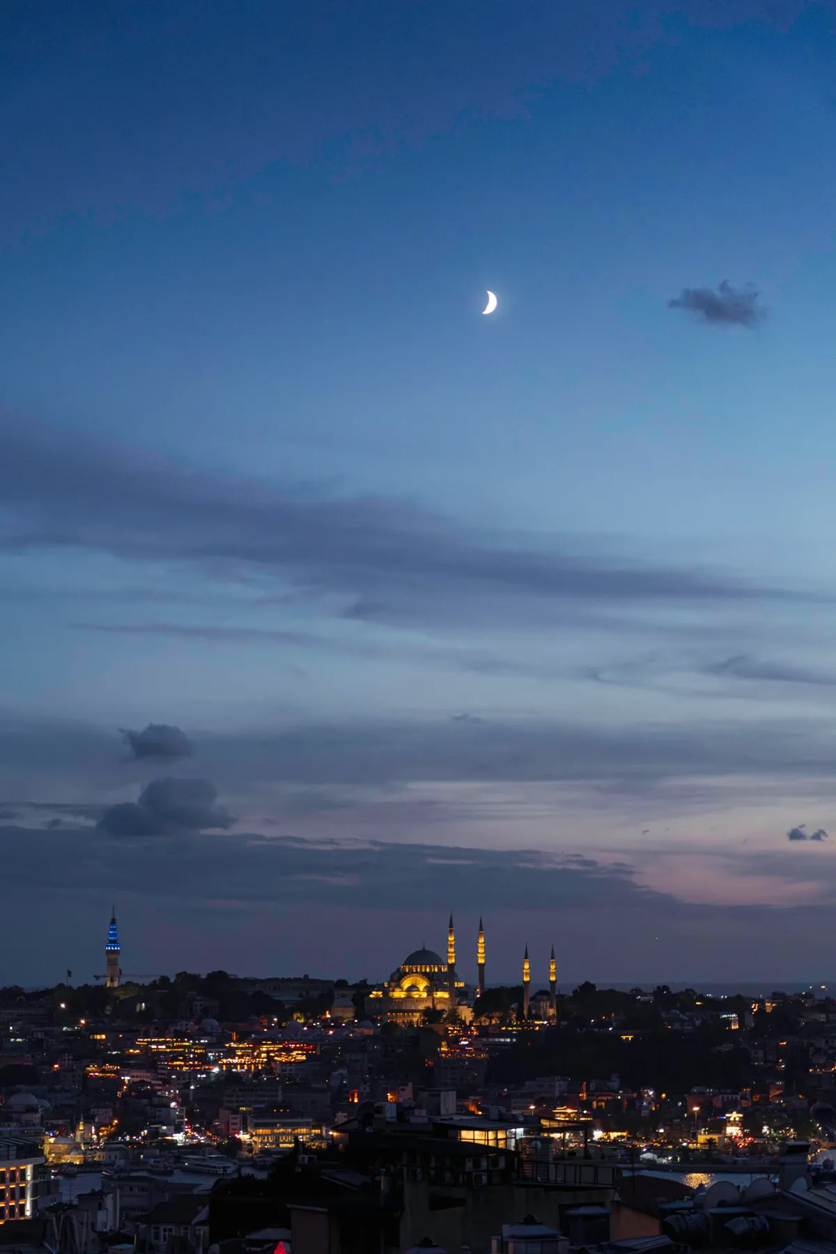A crescent moon over the Istanbul skyline at blue hour, with city lights and a floodlit mosque in the distance.