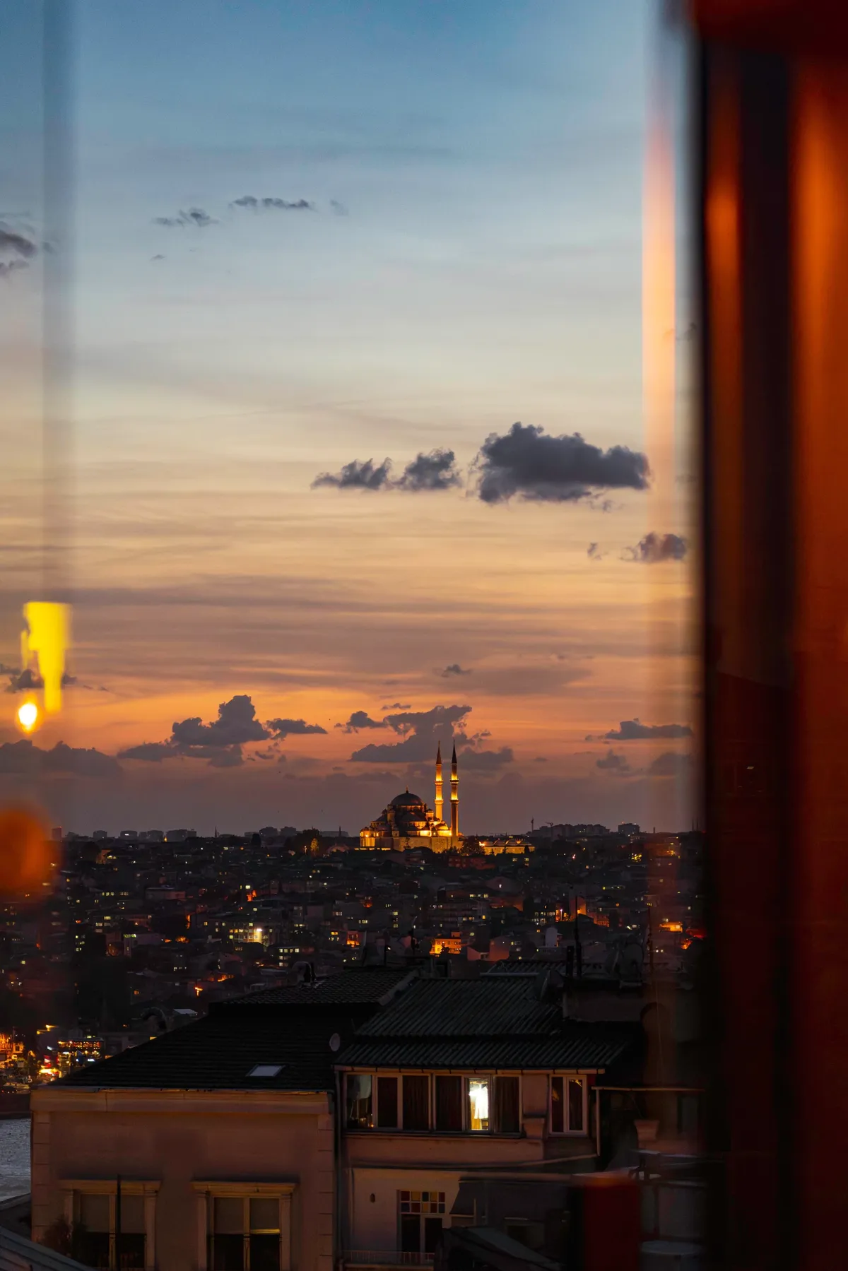 A floodlit mosque with two minarets on the Istanbul skyline at sunset, seen through a window pane with orange reflections.
