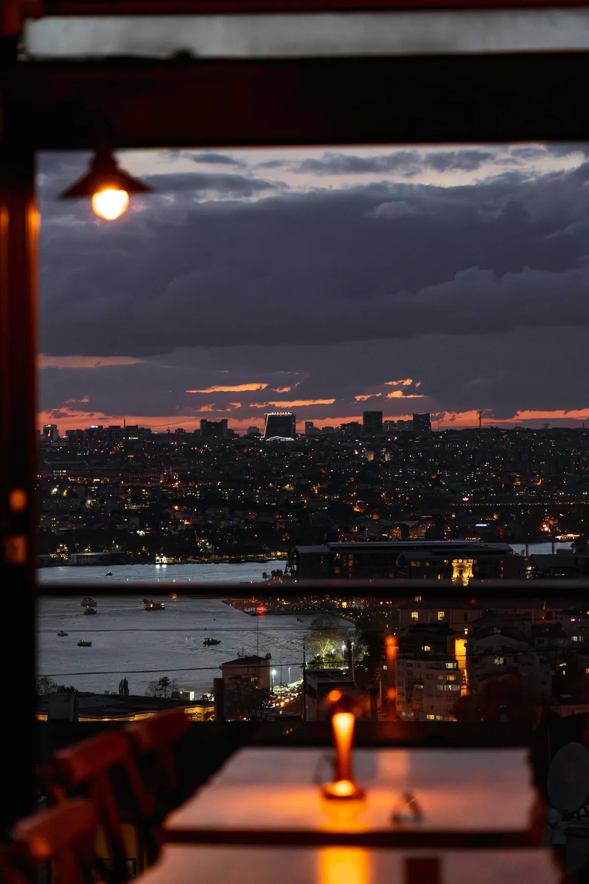 A candle and hanging lamp on a table beside a window overlooking the Golden Horn and Istanbul's lights at dusk.