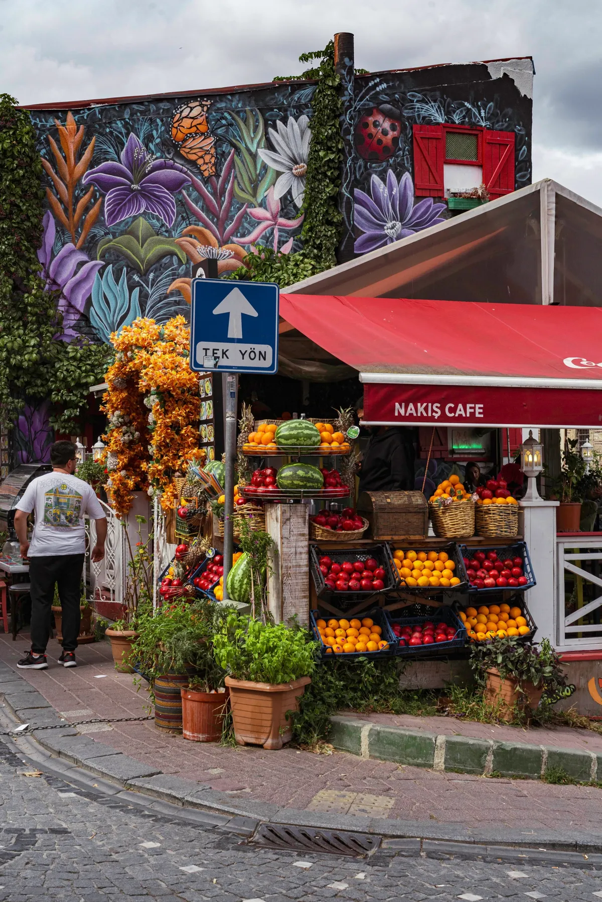 A street corner in Istanbul with a flower mural above a fruit stand, and a man in white walking by.