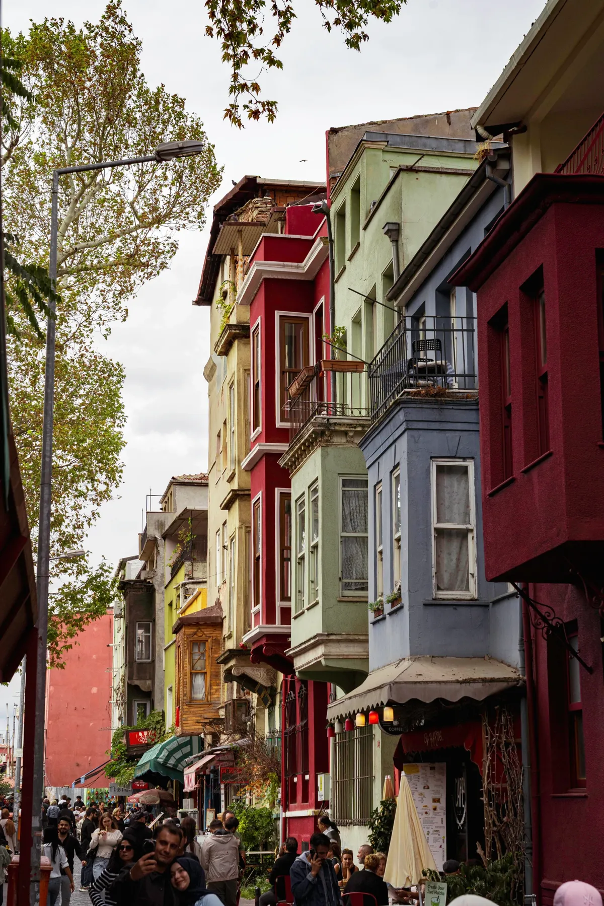 A lane of tall red, blue, green and yellow wooden houses with bay windows, with people walking below in Istanbul.