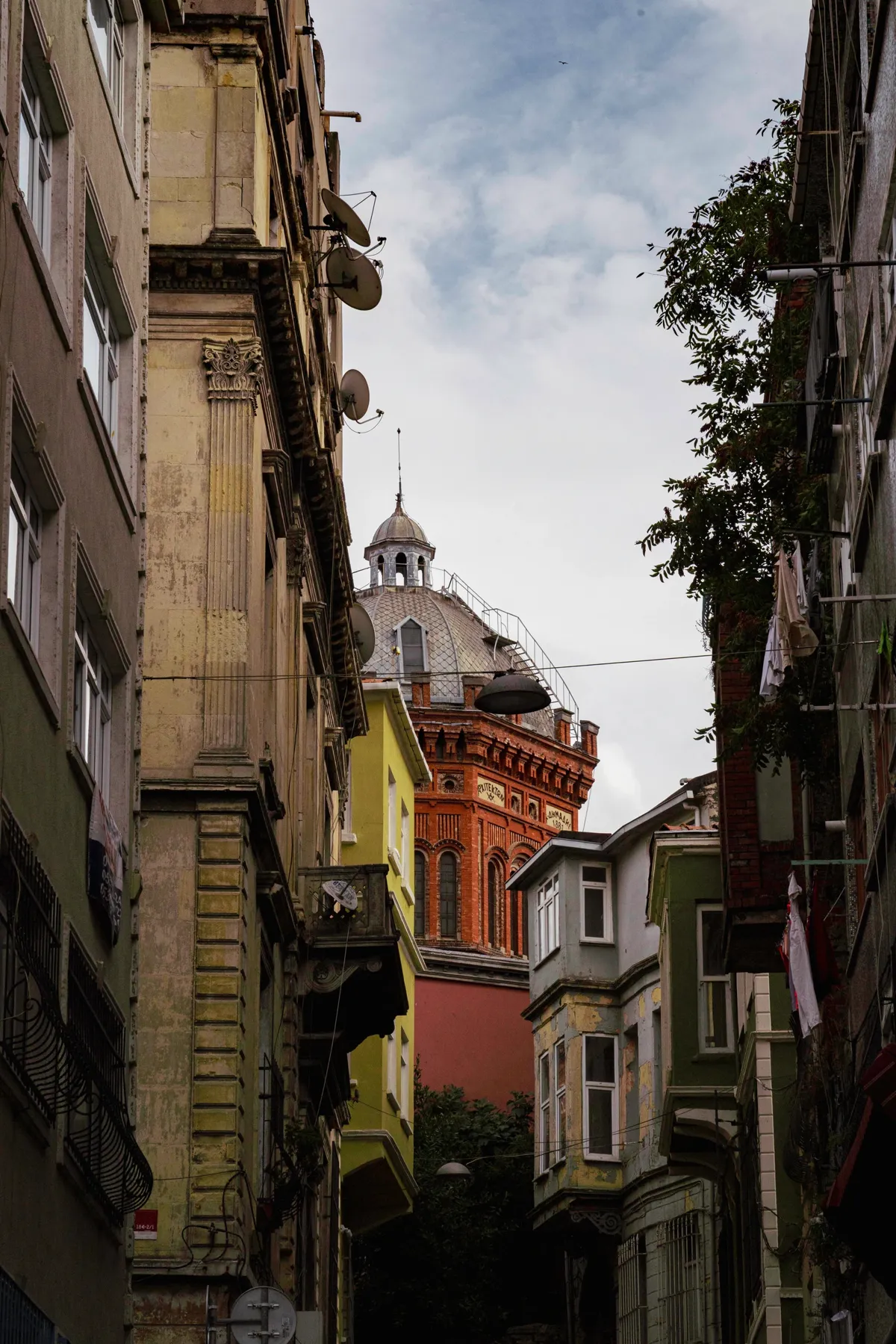 A narrow Istanbul street between worn apartment buildings with satellite dishes, with a red-brick domed building in the distance.