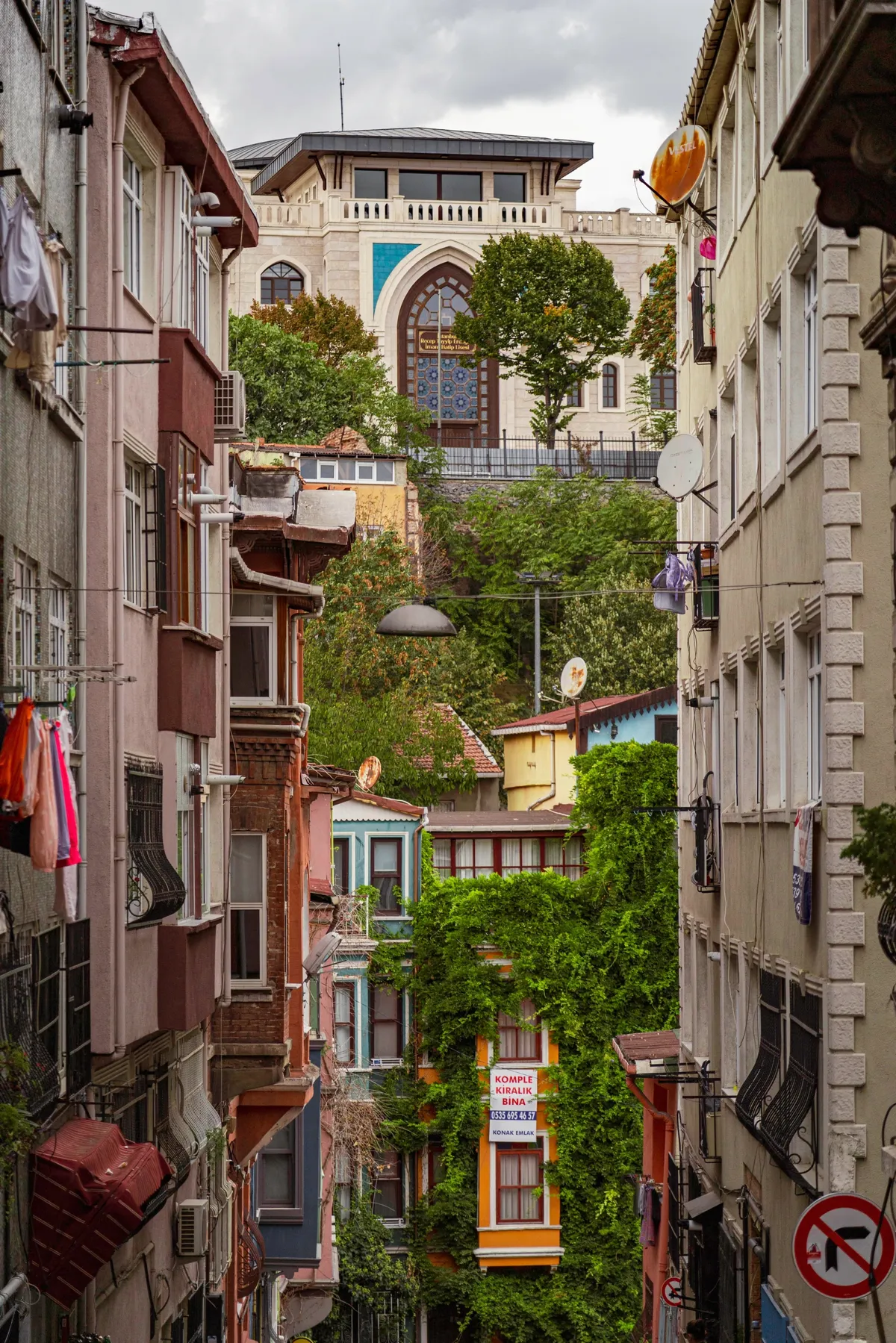 A steep narrow Istanbul street with ivy-covered houses and a large stone building with a tiled window above the rooftops.