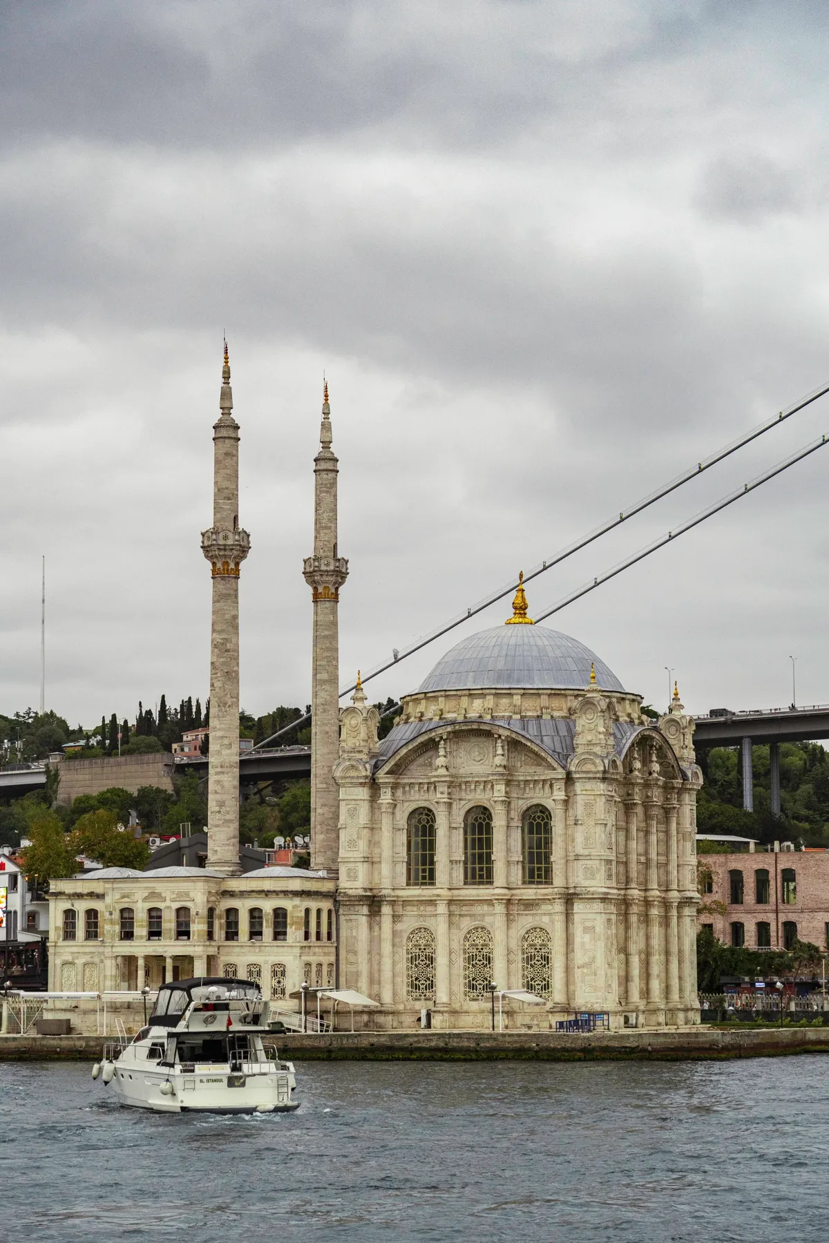 An ornate stone mosque with a dome and two tall minarets on the Bosphorus shore, with a bridge and motorboat.