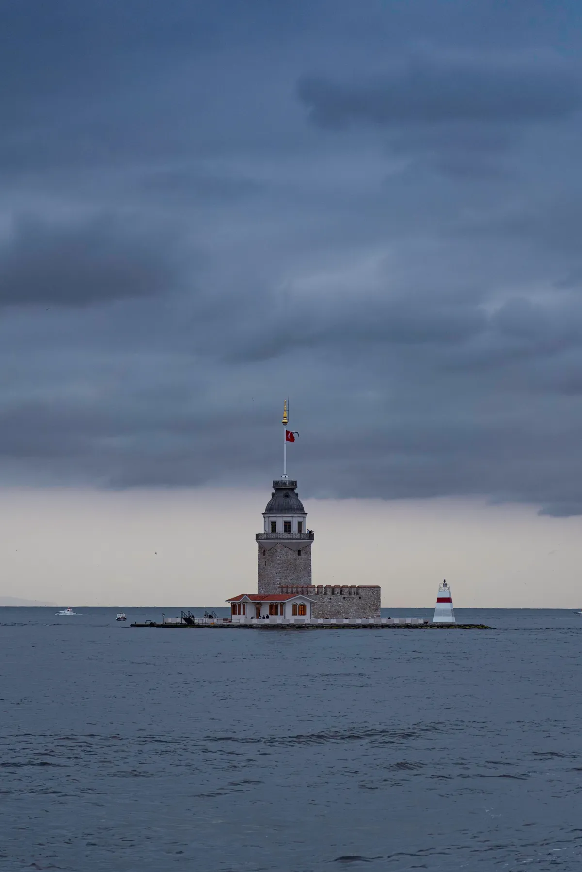 A small stone tower with a flag stands on an islet in the Bosphorus under heavy grey clouds.