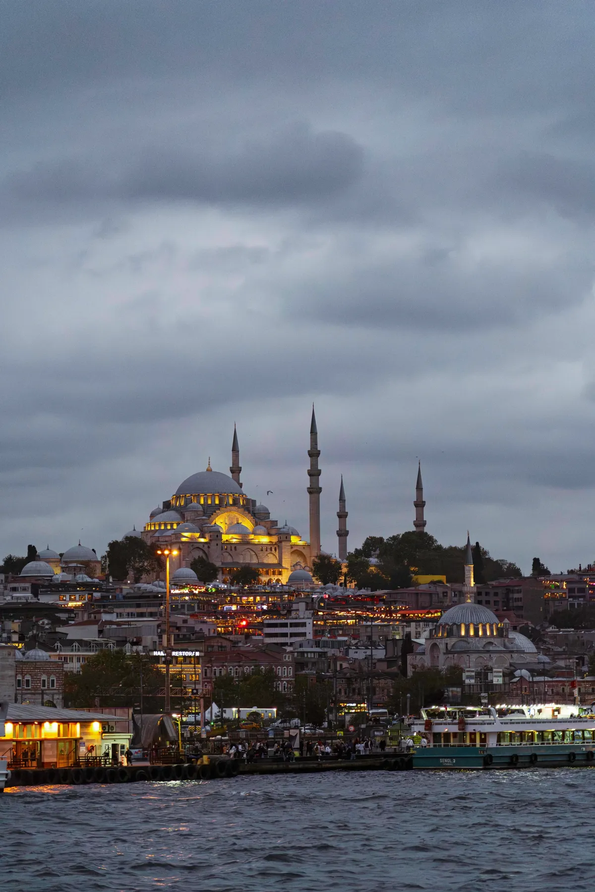 A floodlit mosque with a large dome and slender minarets on a hill above the water at dusk.