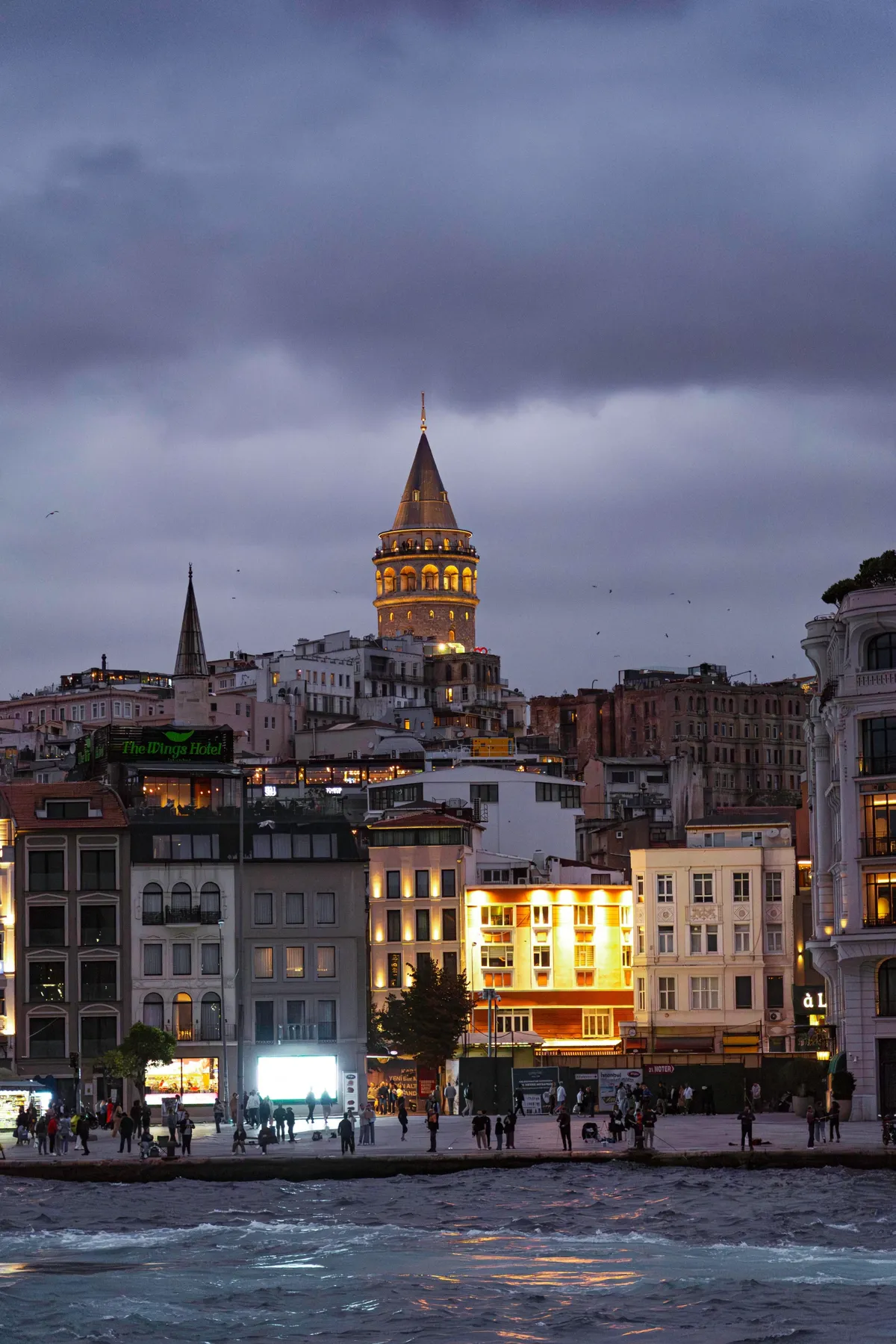 The Galata Tower glows above lit buildings and people along the waterfront in Istanbul under purple dusk clouds.