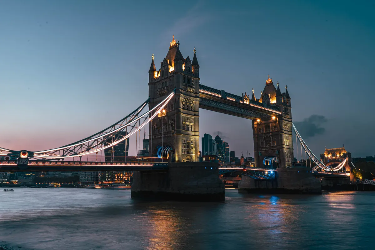 Tower Bridge lit up across the Thames at dusk, with its towers glowing and lights reflected in the water.
