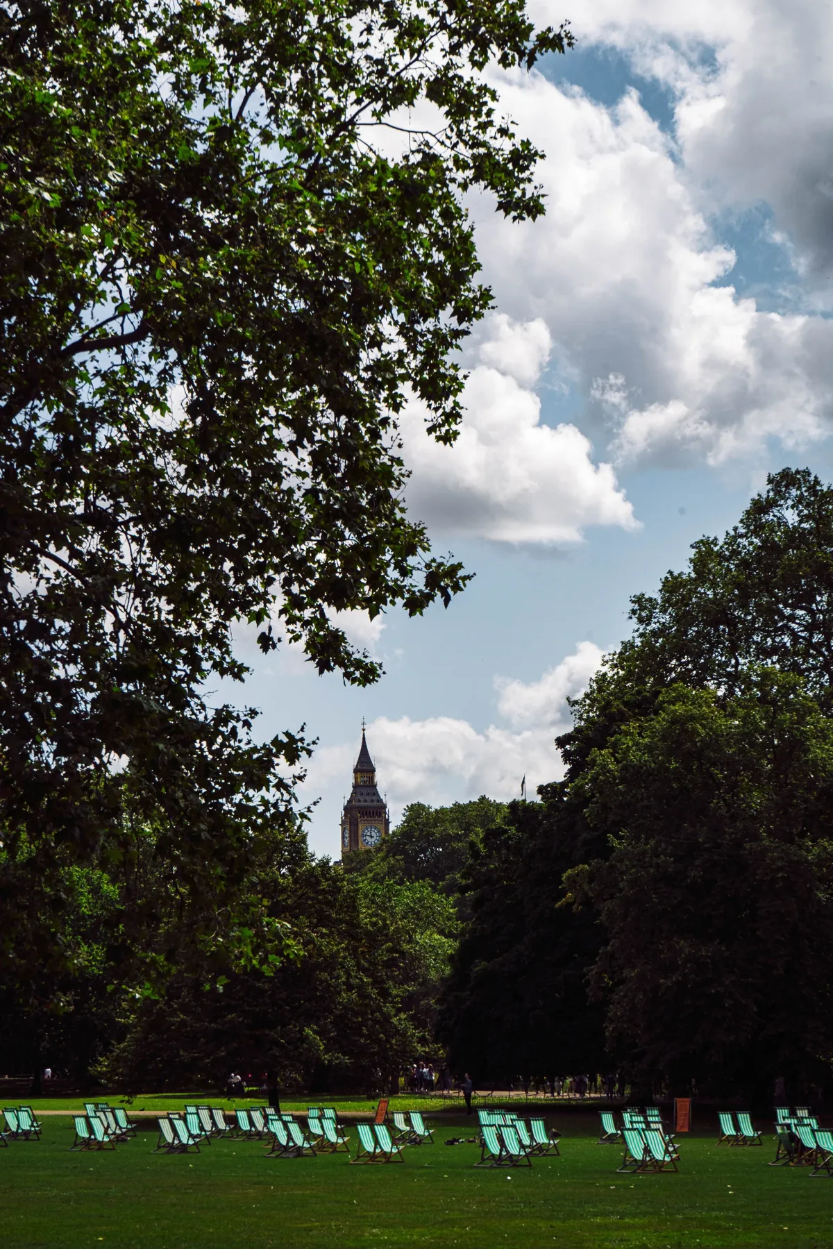 Green deckchairs on a park lawn under large trees, with the Big Ben clock tower rising in the distance.