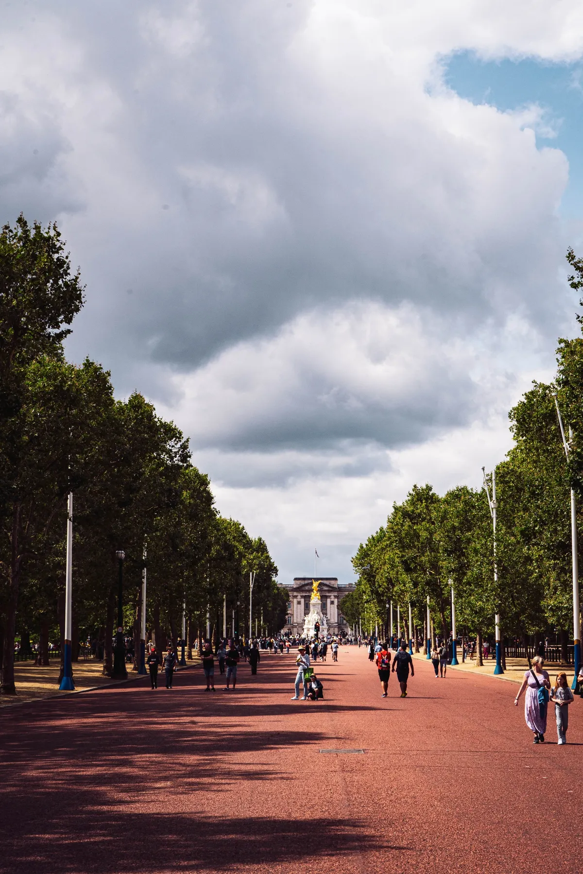A wide red avenue lined with trees leads toward a gold-topped monument and palace, with people walking in the distance.