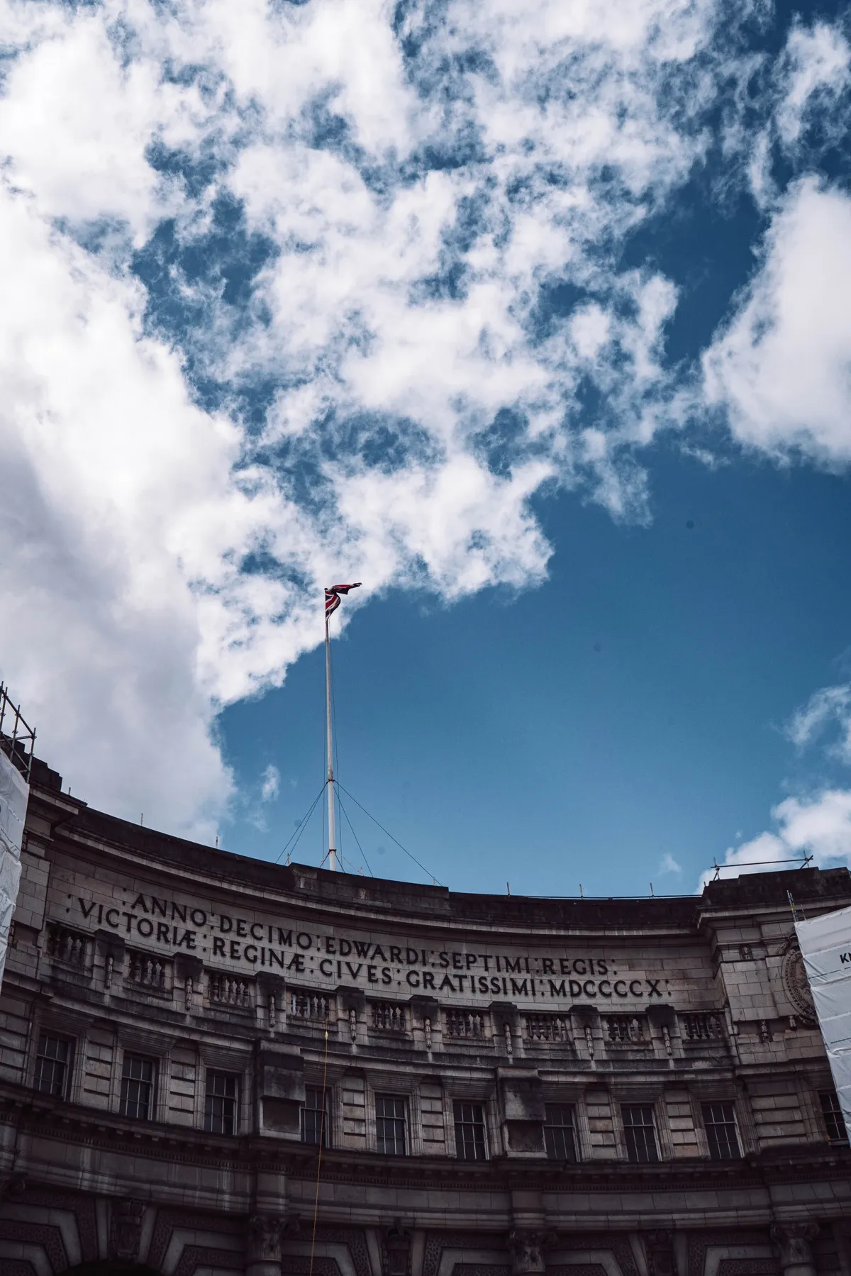 The curved stone facade of Admiralty Arch in London, with a flag on a pole against a blue sky.