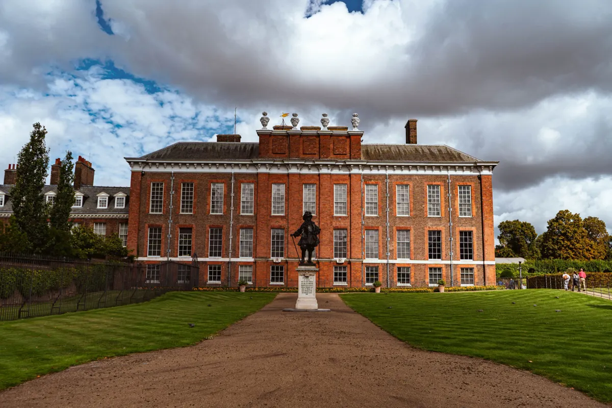 A red-brick palace with a bronze statue on a plinth, seen across lawns under a dramatic cloudy sky.
