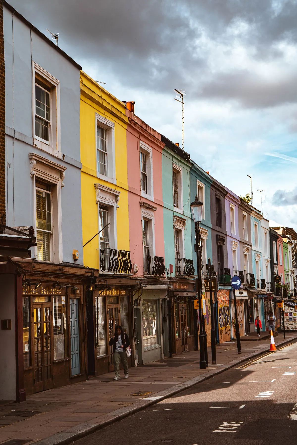 Pastel terraced houses in yellow, pink, blue and green line a London street under cloudy skies, with a person walking.