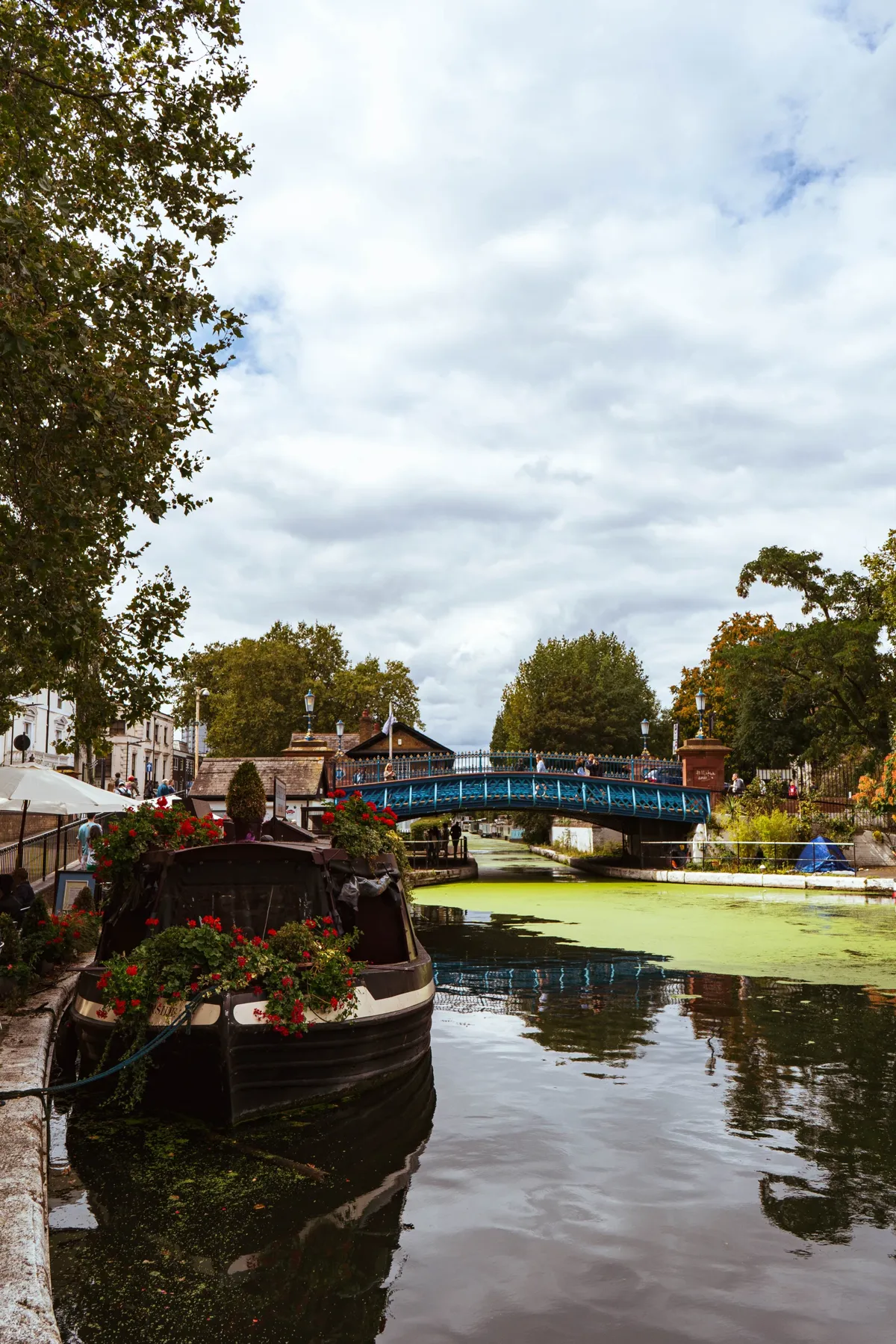 A narrowboat decorated with flowers floats on a canal toward a blue iron bridge, under a cloudy sky.