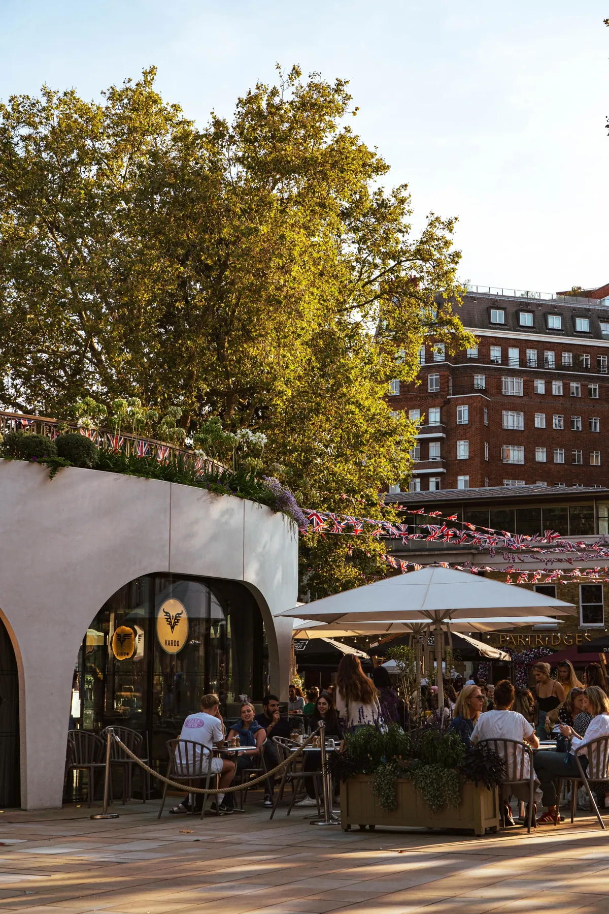 People sit under large parasols at an outdoor cafe beneath tall trees, with bunting and a brick apartment building behind.