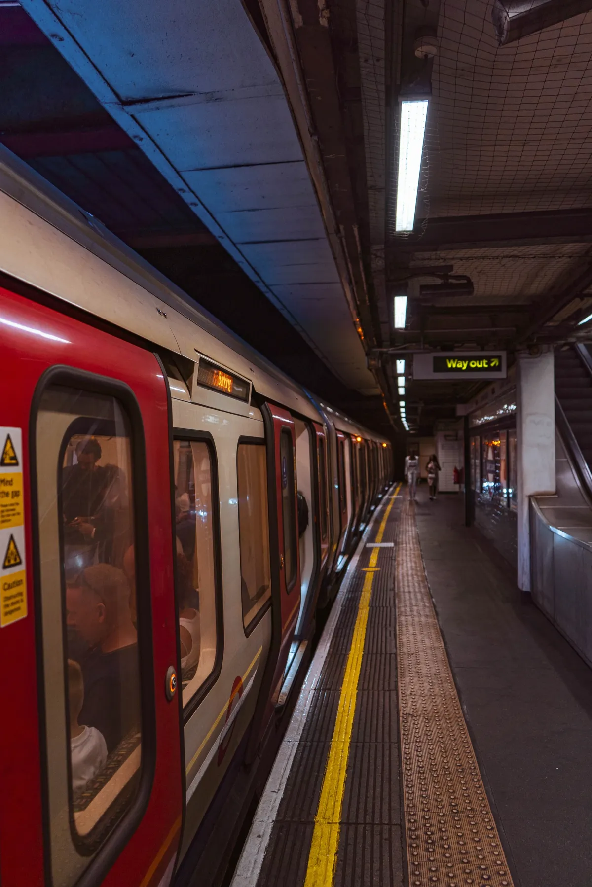 A red London Underground train stands at a platform beside a yellow safety line, with a few passengers far ahead.