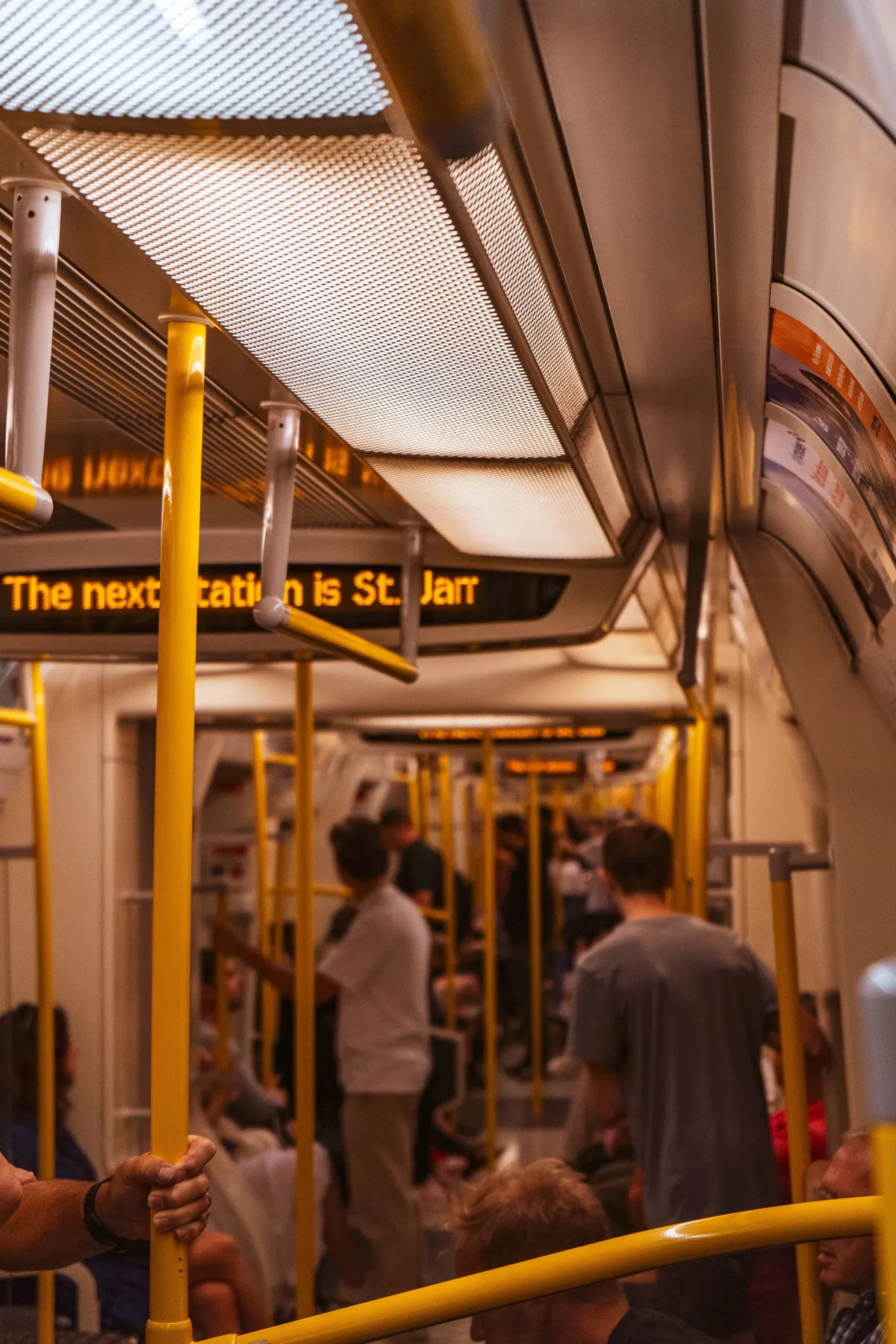 Passengers stand and sit inside a London Underground carriage with yellow handrails and a display announcing the next station.