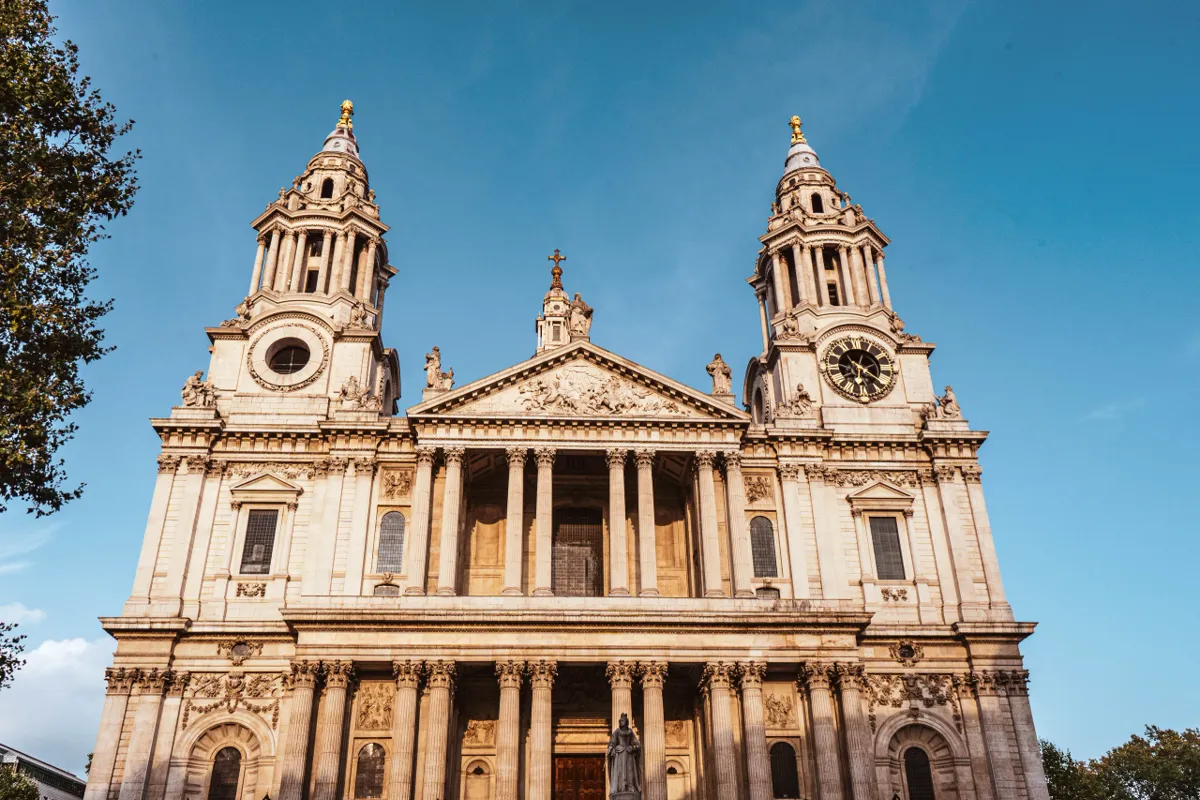 The west front of St Paul's Cathedral, with twin towers and columned portico in golden light under a blue sky.