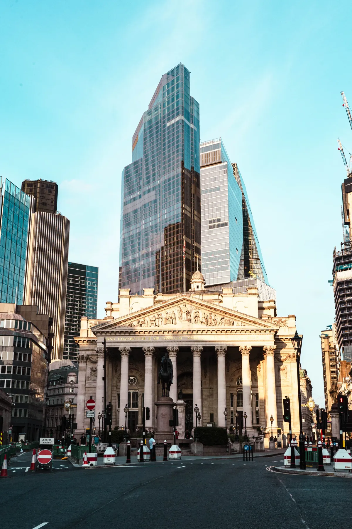 The columned front of the Royal Exchange in London, with glass skyscrapers rising behind it under a blue sky.