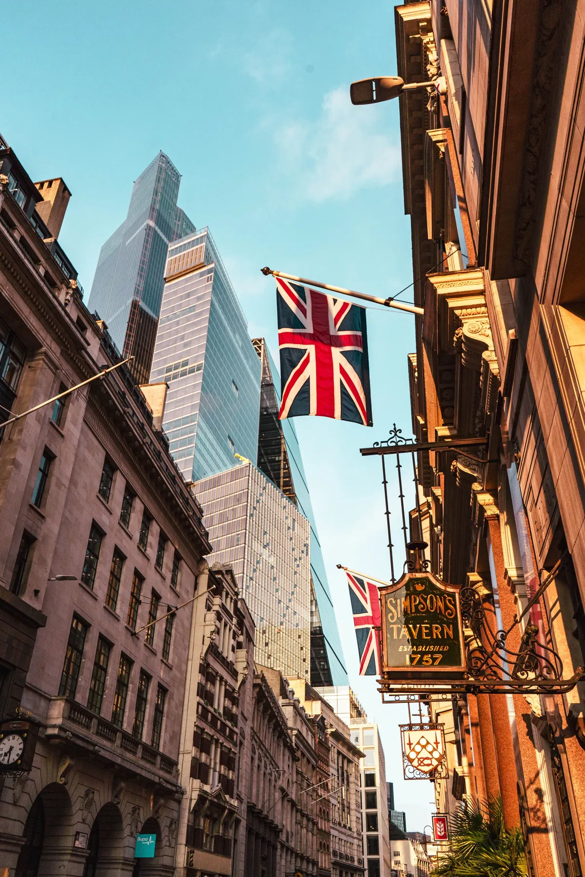 A Union Jack flag and a Simpson's Tavern sign hang over a City of London street, with glass towers above.