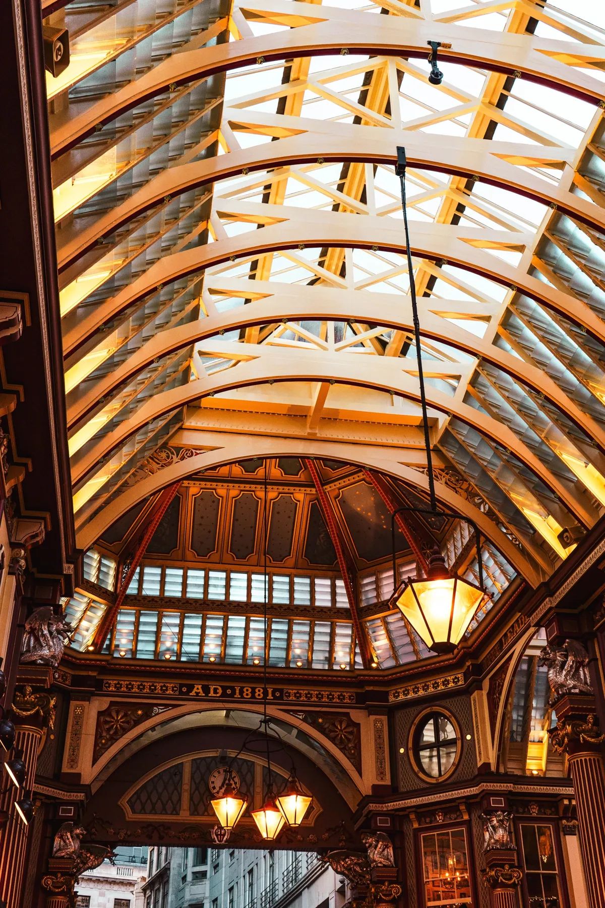 Looking up at the arched glass and iron roof of Leadenhall Market, with ornate painted details and glowing lanterns.