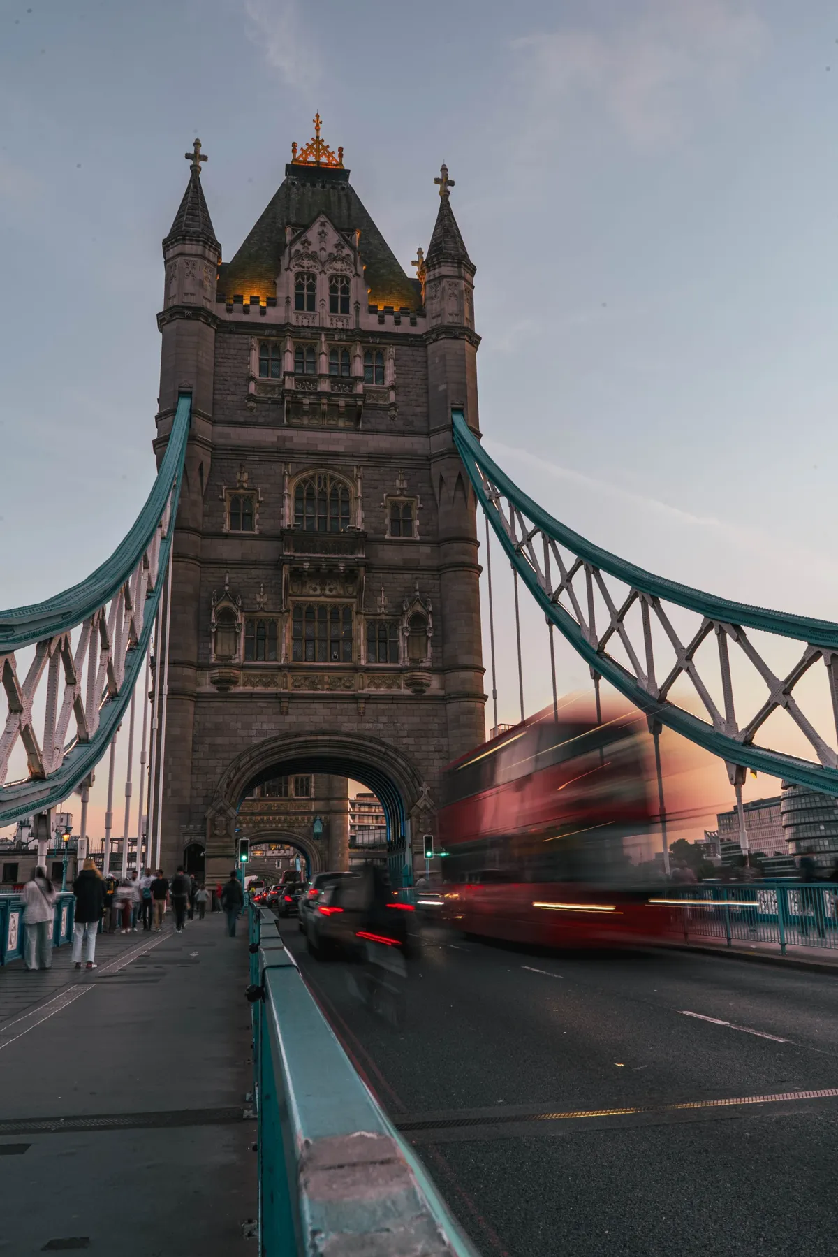 A tower of Tower Bridge in London at dusk, with a blurred red bus crossing the bridge.