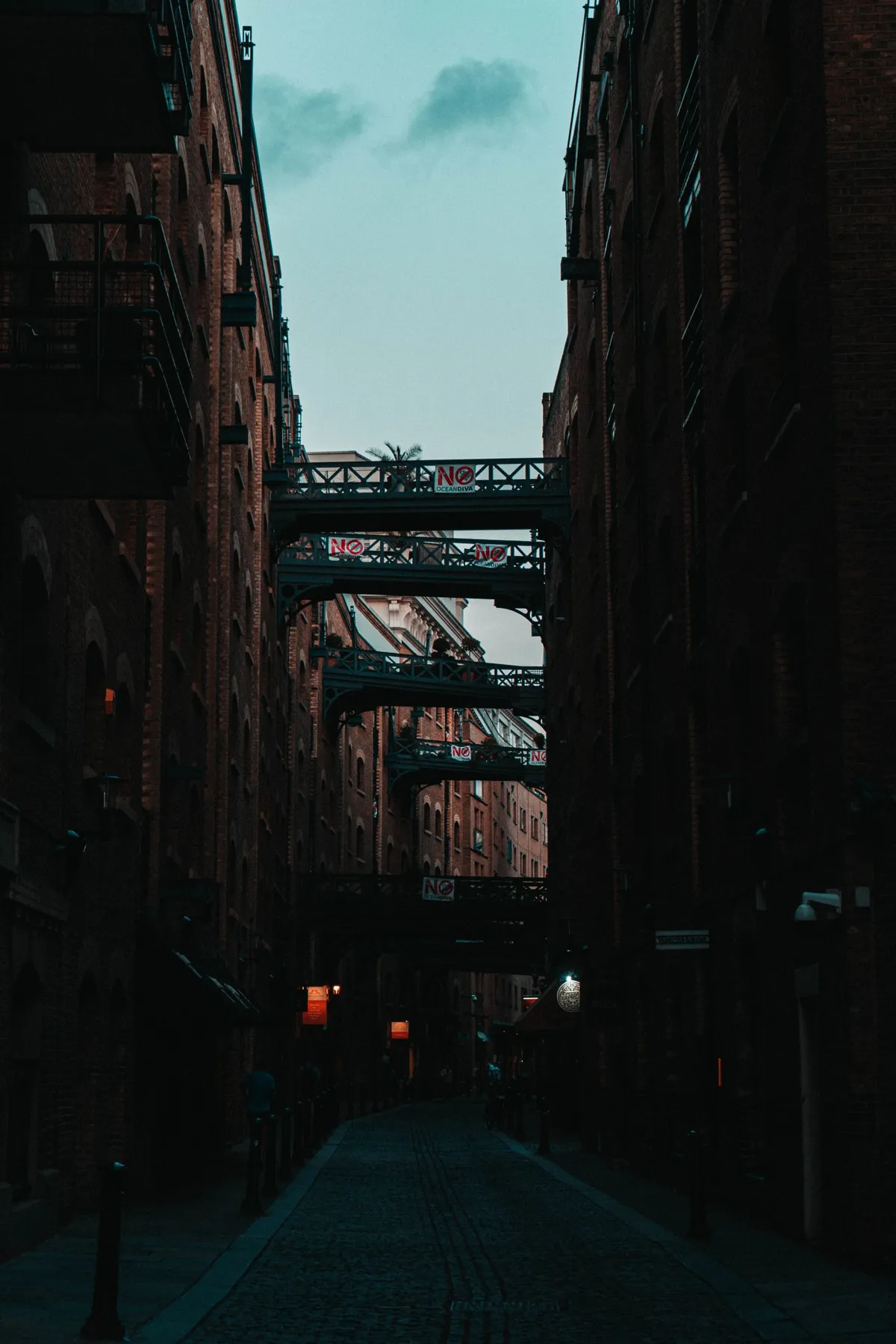 A cobbled lane between dark brick warehouses in London, crossed by iron walkways high overhead at dusk.