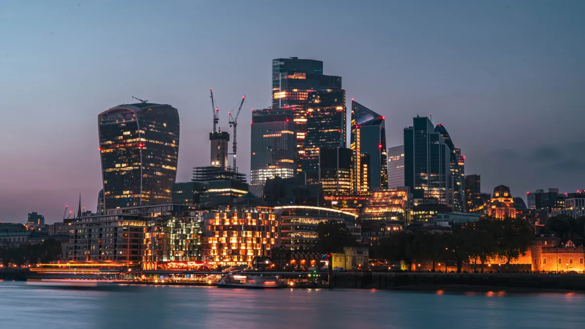 Glass skyscrapers of the City of London glowing at dusk across the Thames, with lit buildings on the waterfront.