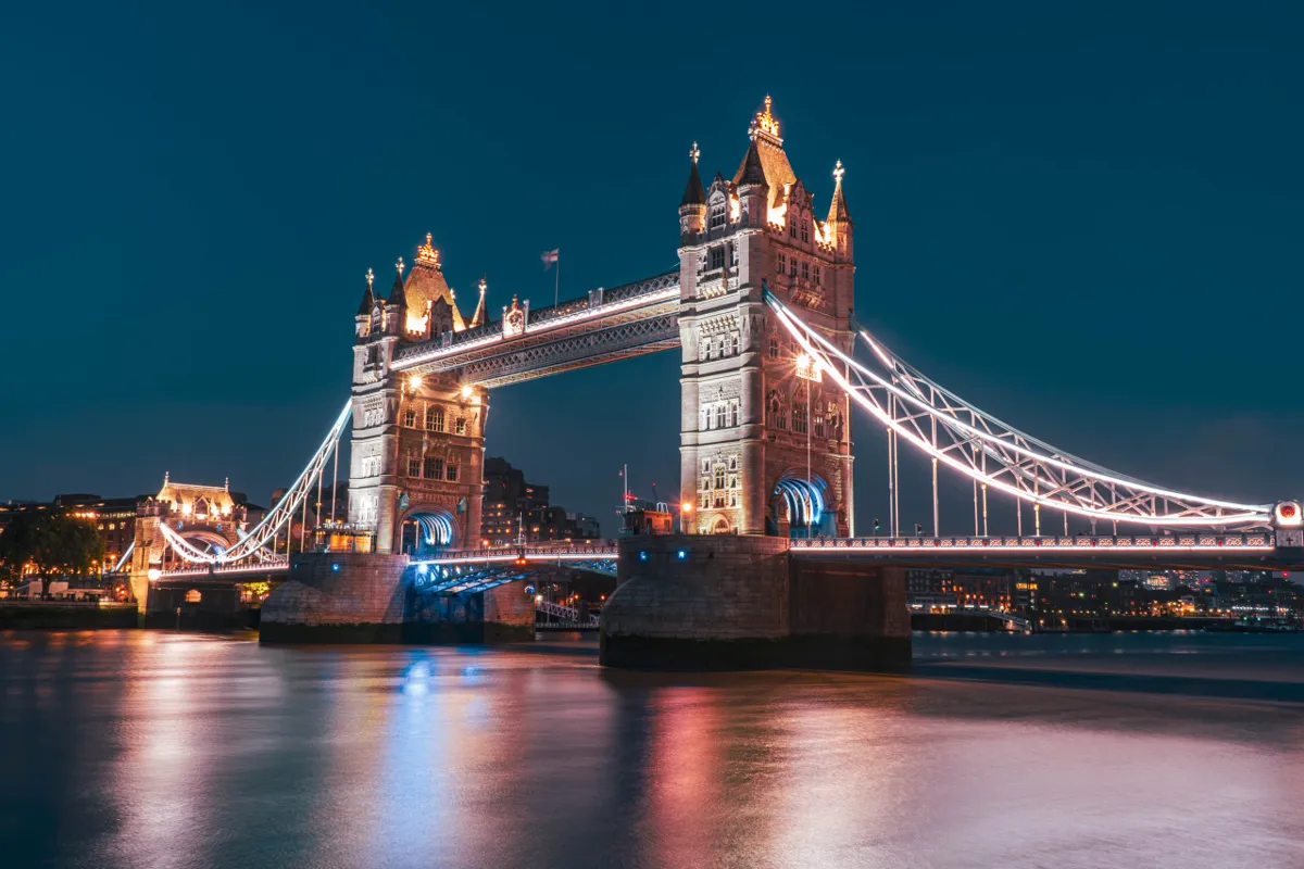 Tower Bridge illuminated at night, its blue-lit arches and white-lit suspension cables reflected in the Thames.