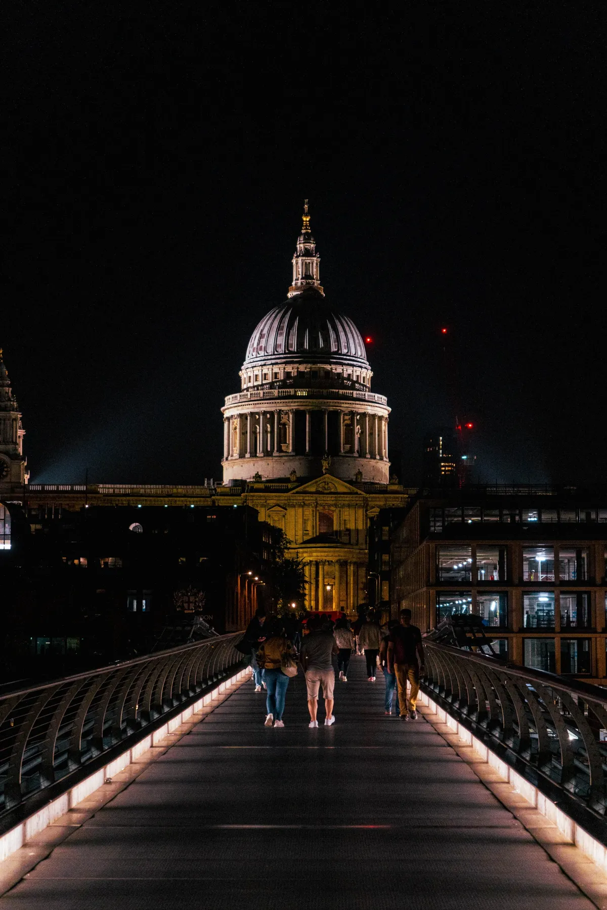 People walk across the Millennium Bridge at night toward the lit dome of St Paul's Cathedral in London.