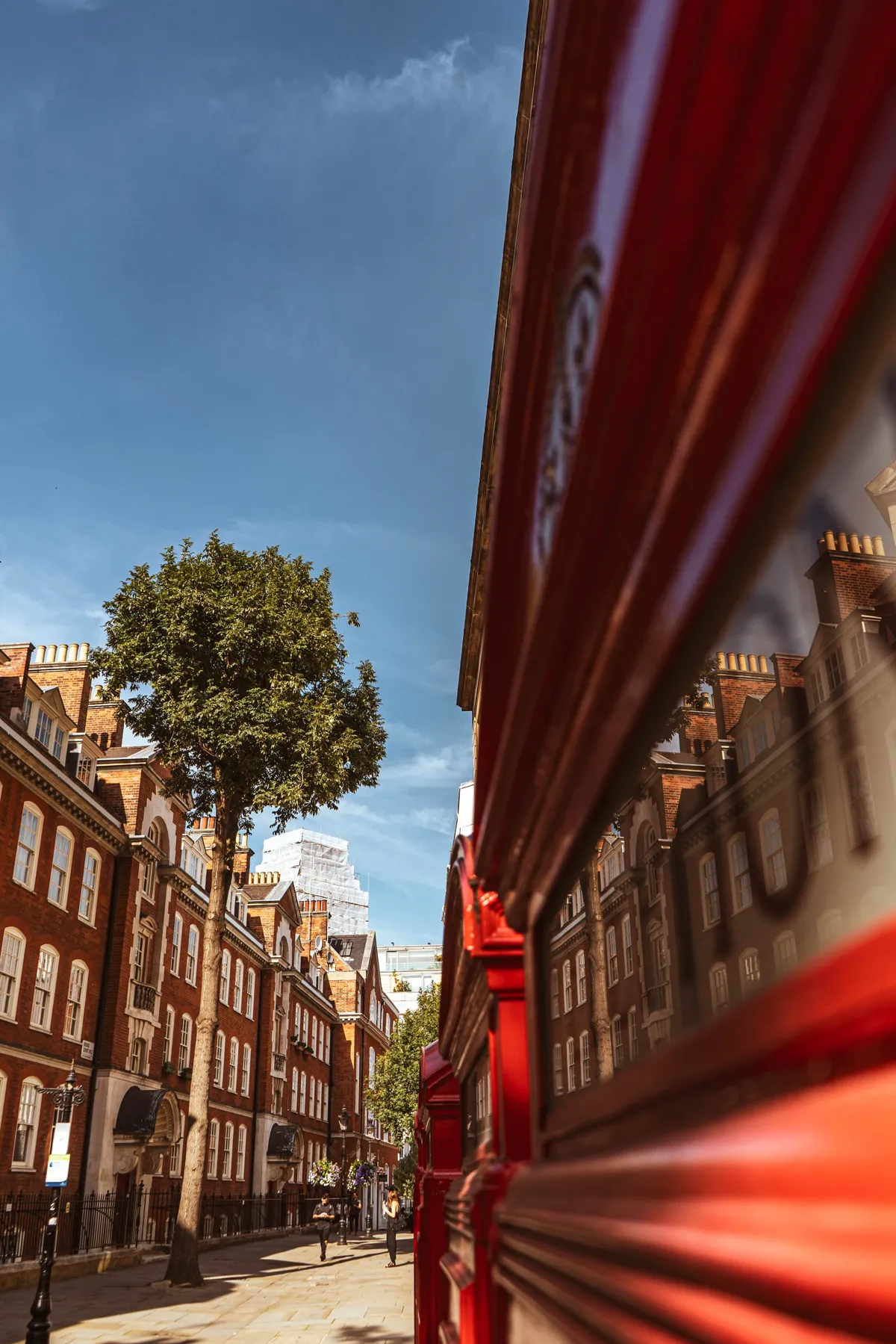 A red telephone box reflects brick buildings on a sunlit London street, with a tree and blue sky beyond.