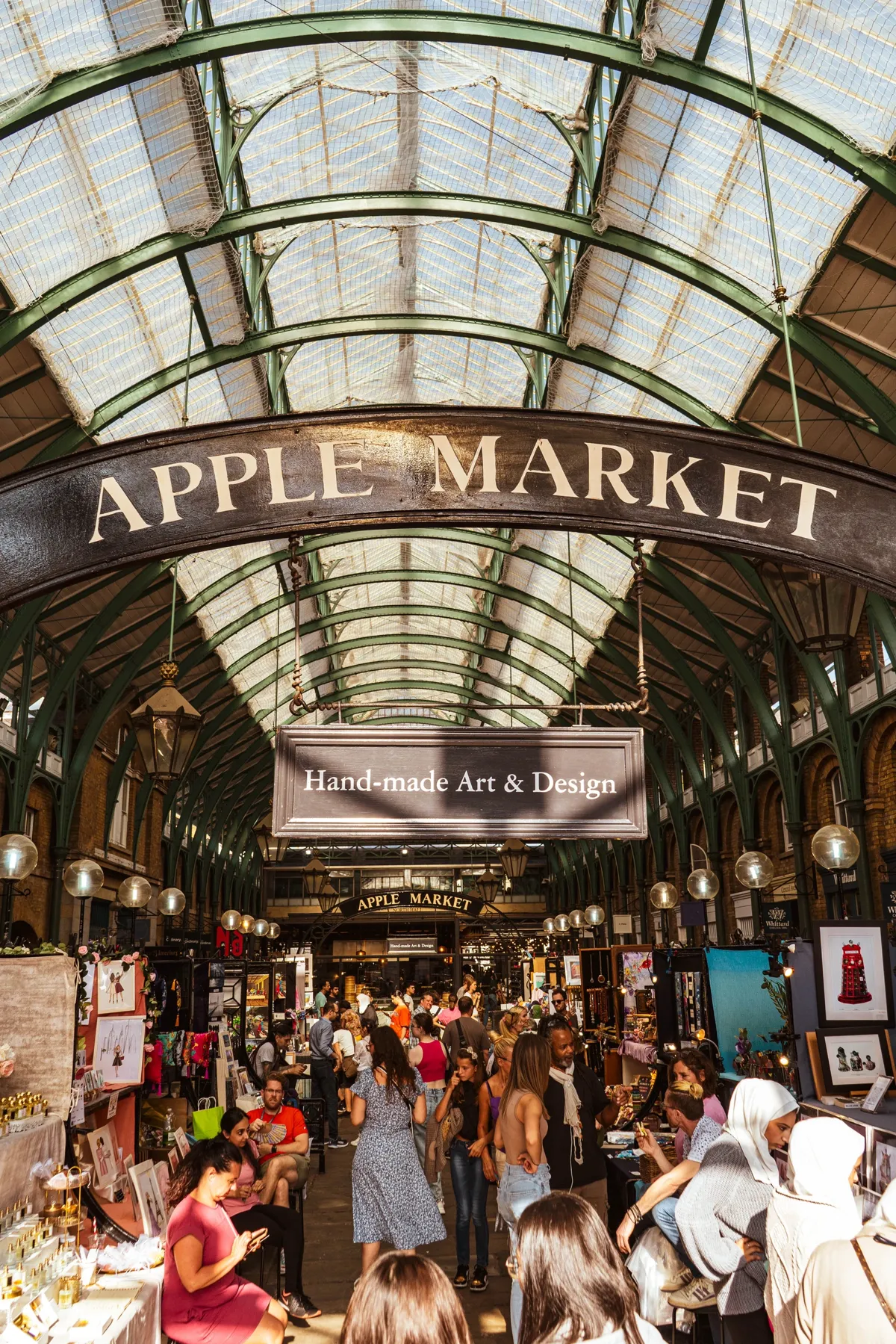 The glass and iron hall of London's Apple Market, with a large sign overhead and crowds browsing craft stalls below.