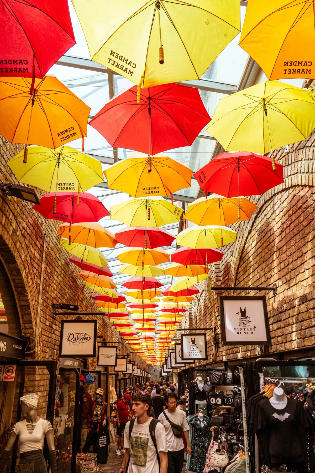 Hundreds of red, yellow and orange umbrellas hang above a brick-walled covered market lane, with shoppers walking below.