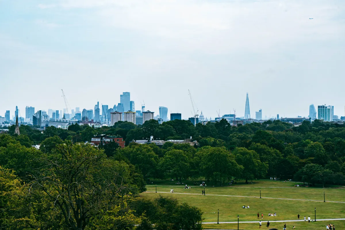 The City of London skyline, with skyscrapers and the Shard, rises over trees and a park lawn where people relax.