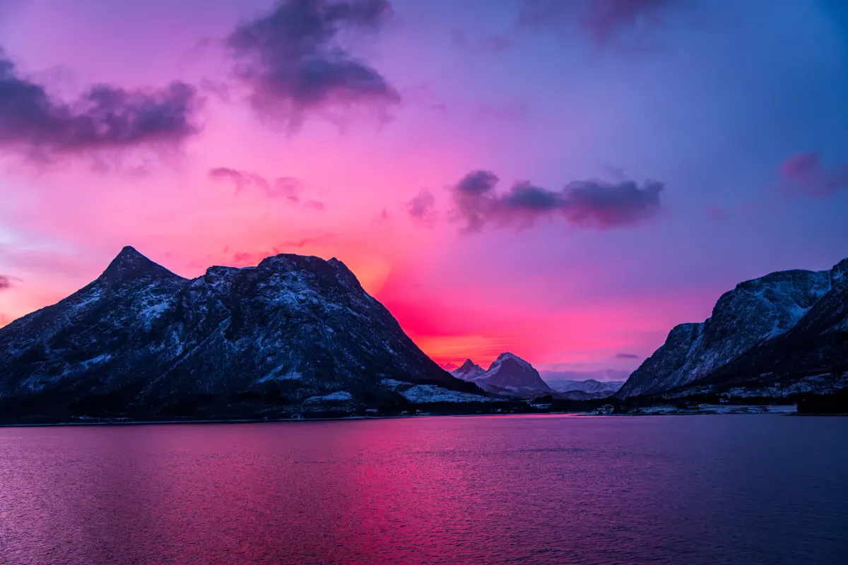 Magenta and violet sunrise sky reflected in a calm fjord between steep, snow-dusted mountains.