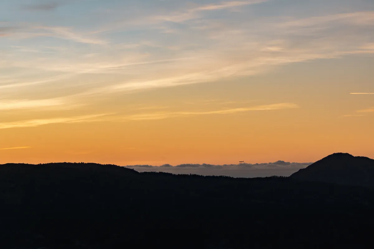 Orange and pink sunset sky over dark silhouetted hills near Bergen, with a tiny aircraft near the horizon.