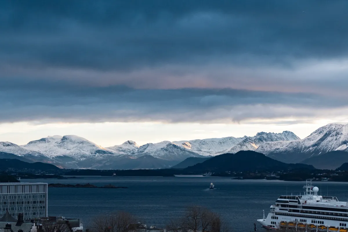 A cruise ship in a fjord harbour with snow-covered mountains and heavy storm clouds behind.