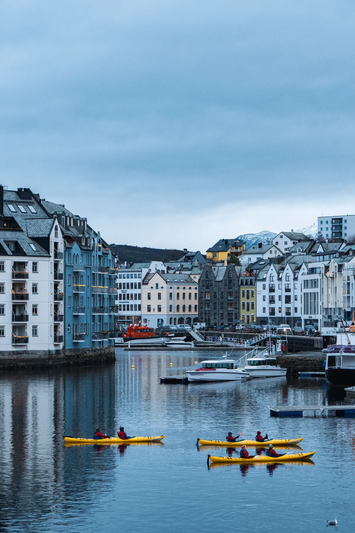 Kayakers in yellow kayaks paddle across a calm harbour canal lined with Art Nouveau buildings and moored boats.