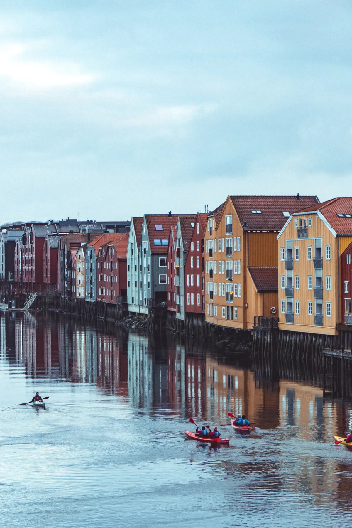 Kayakers paddle on a river beside tall colourful wooden warehouses reflected in the water under a grey sky.