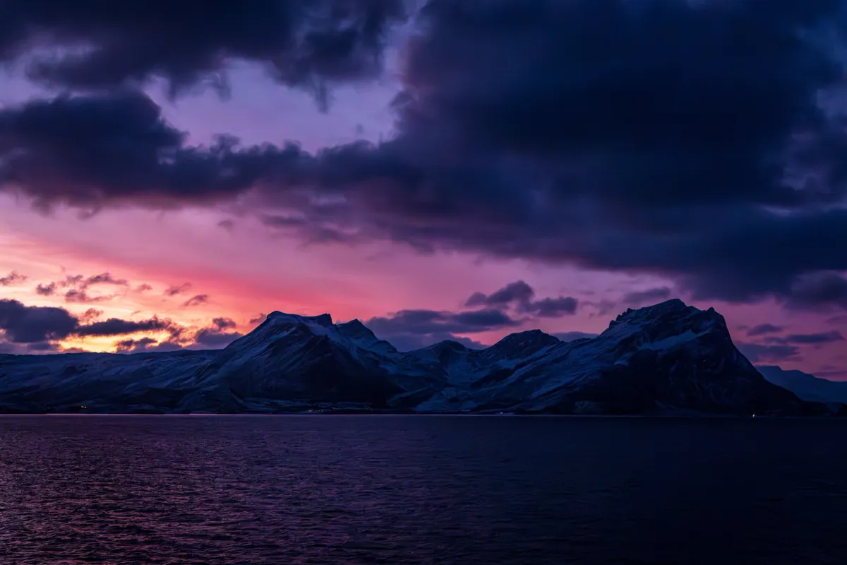 Snow-covered mountains silhouetted against a purple and magenta sunrise sky above dark water, seen from the Norwegian coast.