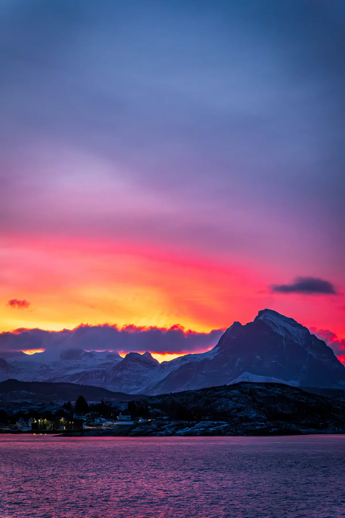 A fiery pink and orange sky glows behind a snowy peak over a dark fjord, with a few lit houses.