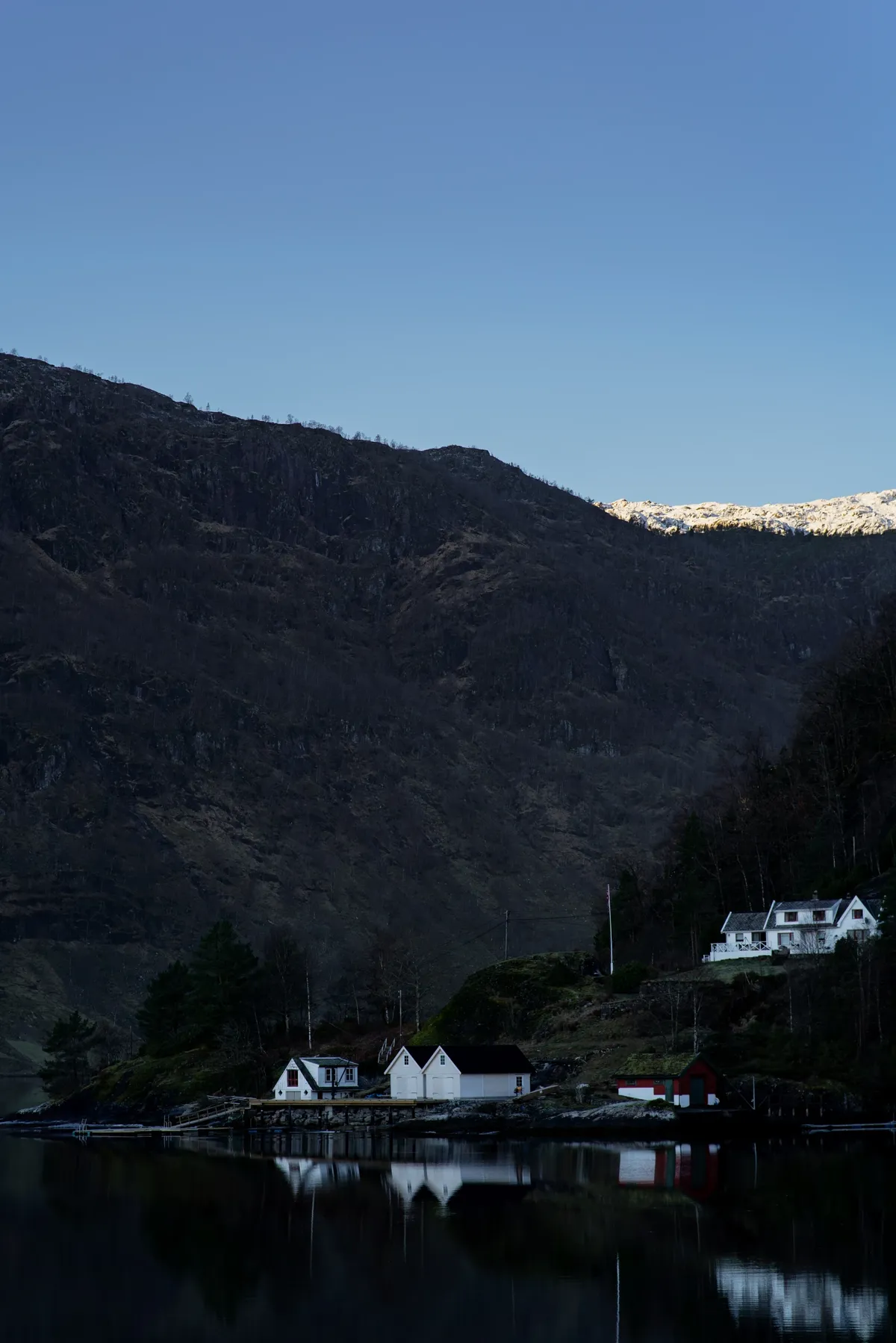 White houses and boathouses on a dark fjord shore with reflections, under a snow-capped ridge near Bergen at dusk.