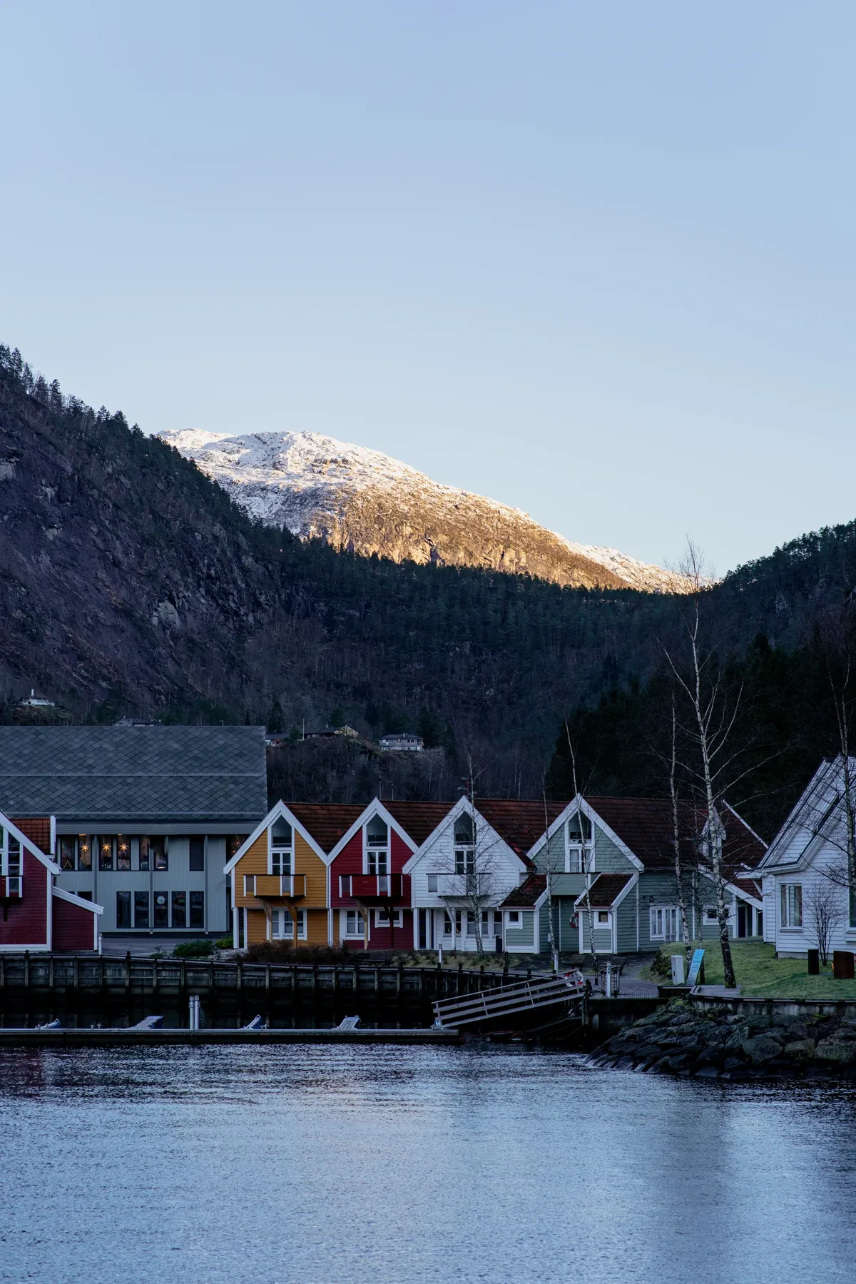 Colourful wooden houses along the water with a snow-dusted mountain behind them, in soft evening light near Bergen.