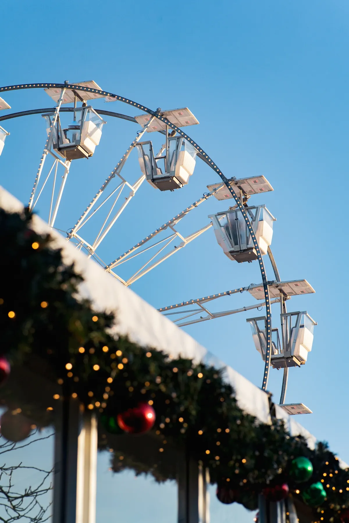 A white Ferris wheel against a blue sky above a market stall roof decorated with Christmas garland and baubles.
