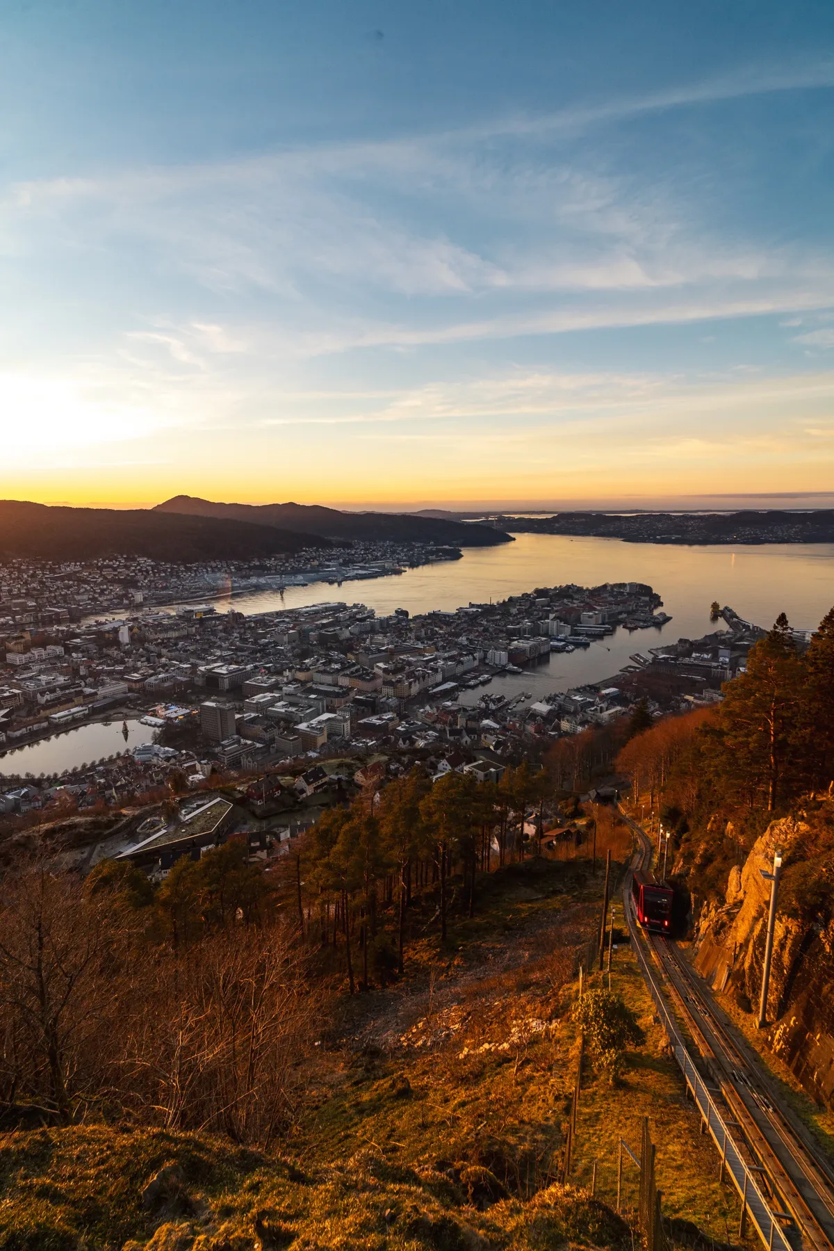Sunset over Bergen and its fjord seen from the hill, with a red funicular car on the track at right.
