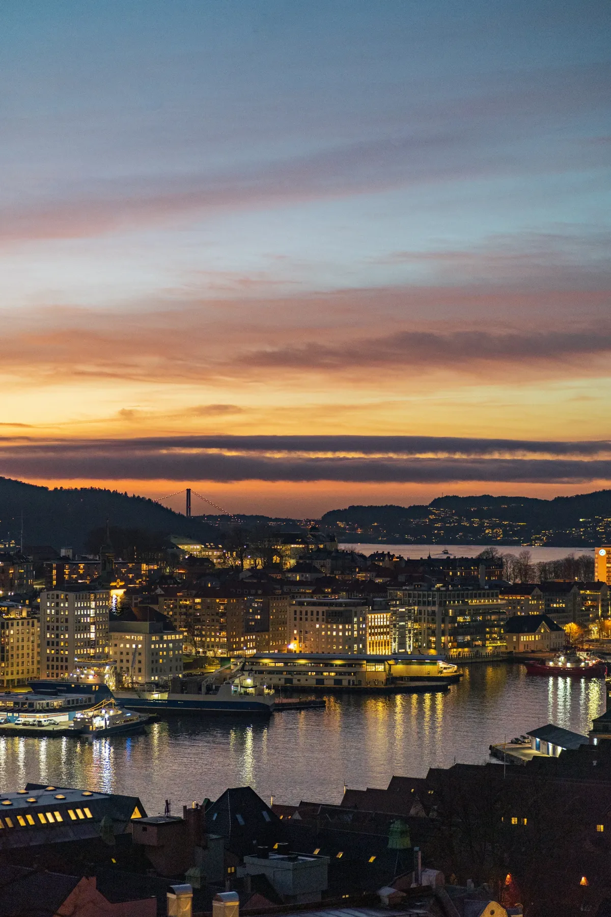 Bergen harbour at dusk with lit buildings reflected in the water and orange clouds above a distant bridge.