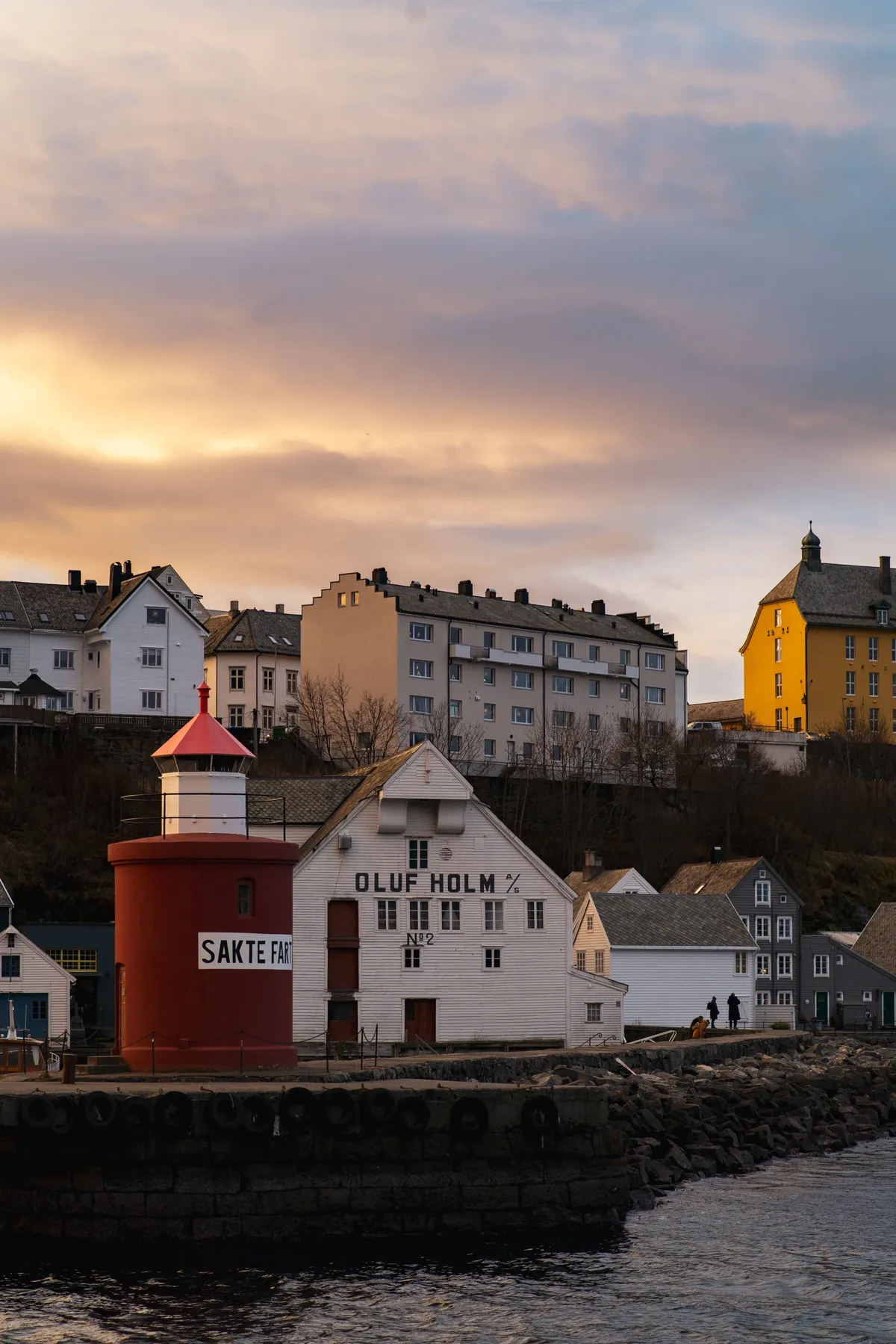 A red and white harbour lighthouse beside old warehouses and hillside houses under a golden sunset sky.