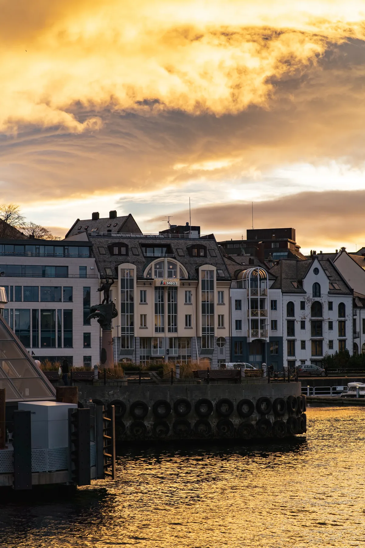 Harbourfront buildings and a small monument beneath dramatic golden clouds at sunset, with warm light on the water.