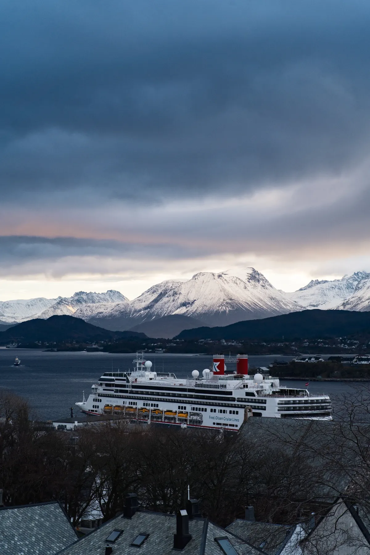 A white cruise ship moored in front of snowy mountains under dark, moody clouds, seen over rooftops.